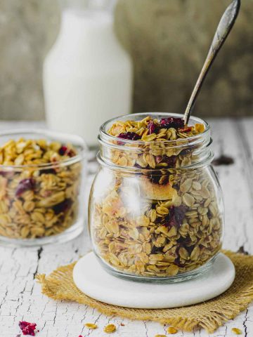 A jar filled with homemade granola with a spoon sticking out of the jar and a small bottle of milk in the background