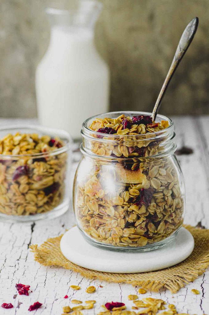 A jar filled with homemade granola with a spoon sticking out of the jar and a small bottle of milk in the background