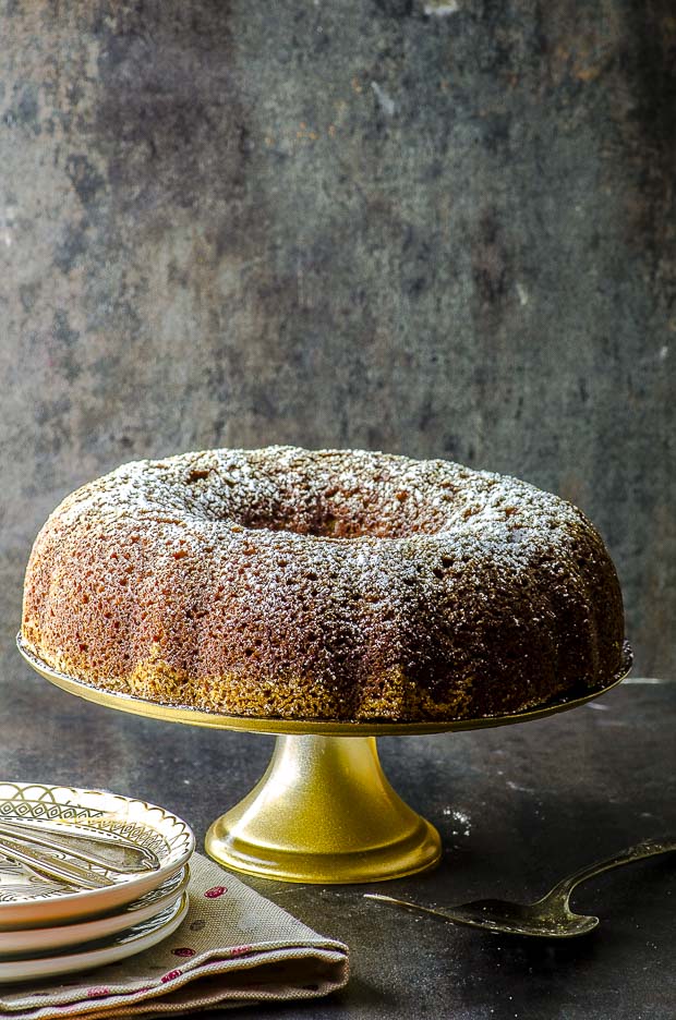 A whole passover walnut cake on a cake stand with a gold base. on the table there is also a partial view of 3 plates staked under a napkin and 3 spoons on top of the plate.