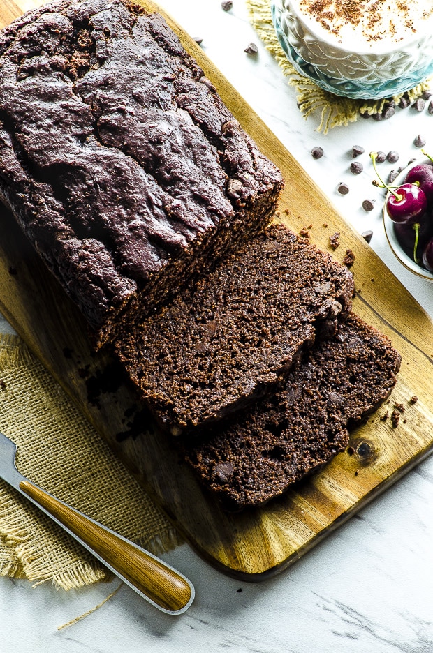 Vegan Chocolate Cake sliced on a wooden cutting board with a white and blue capuccino mug and a small bowl of fresh cherries