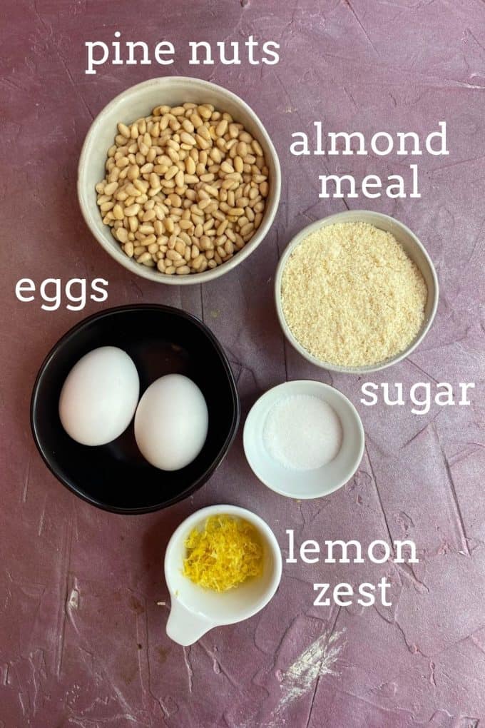 An overhead view of the ingredients to make pignoli cookies; pine nuts, almond meal, eggs, sugar, lemon zest