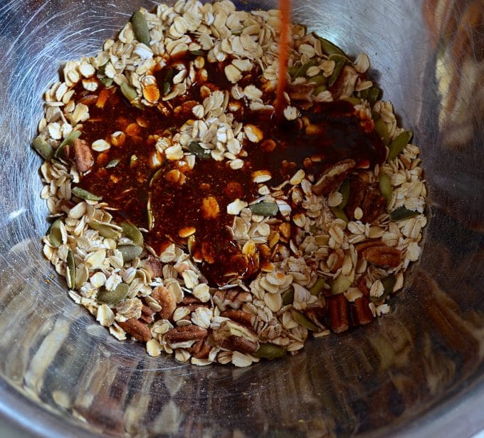 Pouring a mix of spices, oil, maple syrup to a bowl of oats, pecans and pumpkin seeds to make savory granola