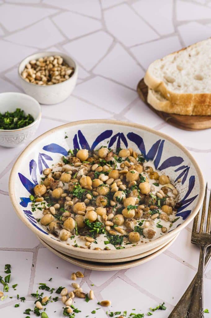 A plate with musabaha next to some bread and two small bowl with pine nuts and parsley