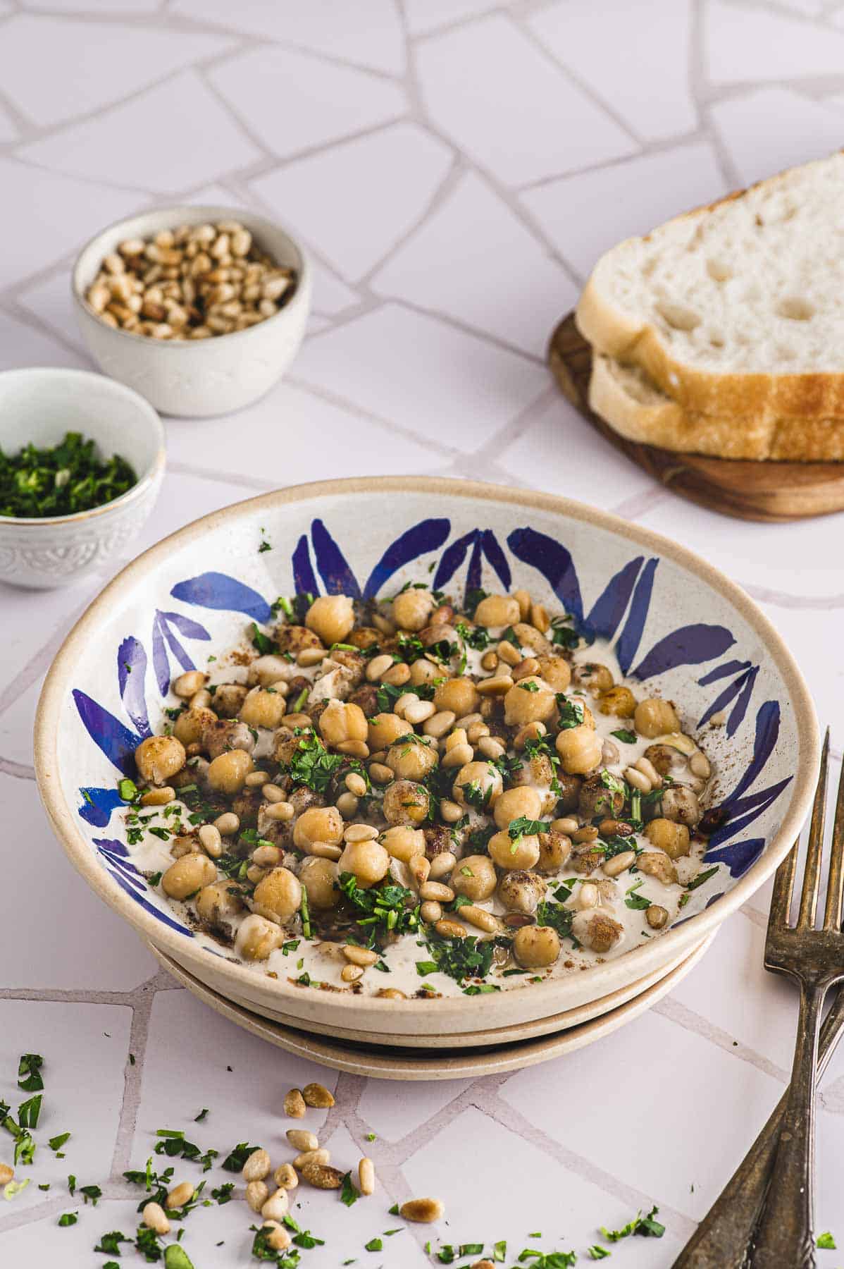 A plate with musabaha next to some bread and two small bowl with pine nuts and parsley