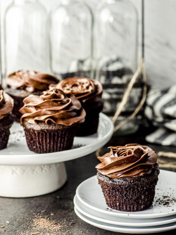 Side view of four chocolate cupcakes with chocolate frosting on a white cake stand and one chocolate cupcake on a stack of white plates