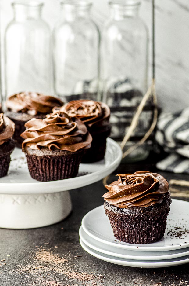 Side view of four chocolate cupcakes with chocolate frosting on a white cake stand and one chocolate cupcake on a stack of white plates