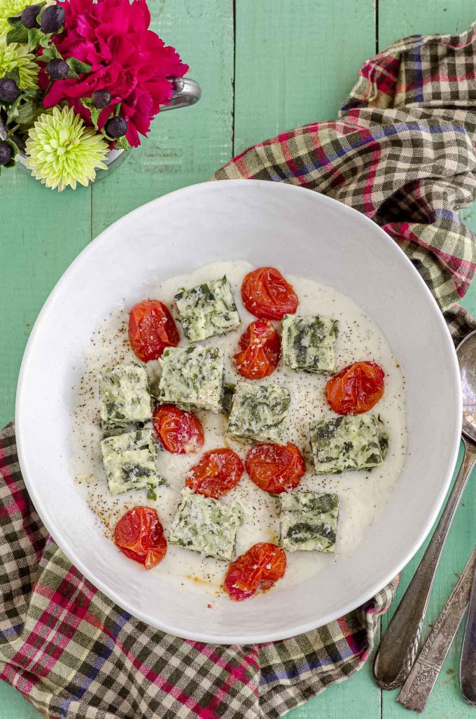Overhead view of a white bowl with spinach semolina gnocchi square over a béchamel sauce and roasted tomatoes