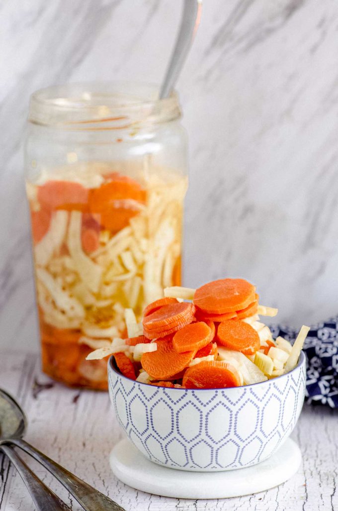 A white and blue bowl filled with pickled carrots and fennel with a jar with the same pickled veggies in the background