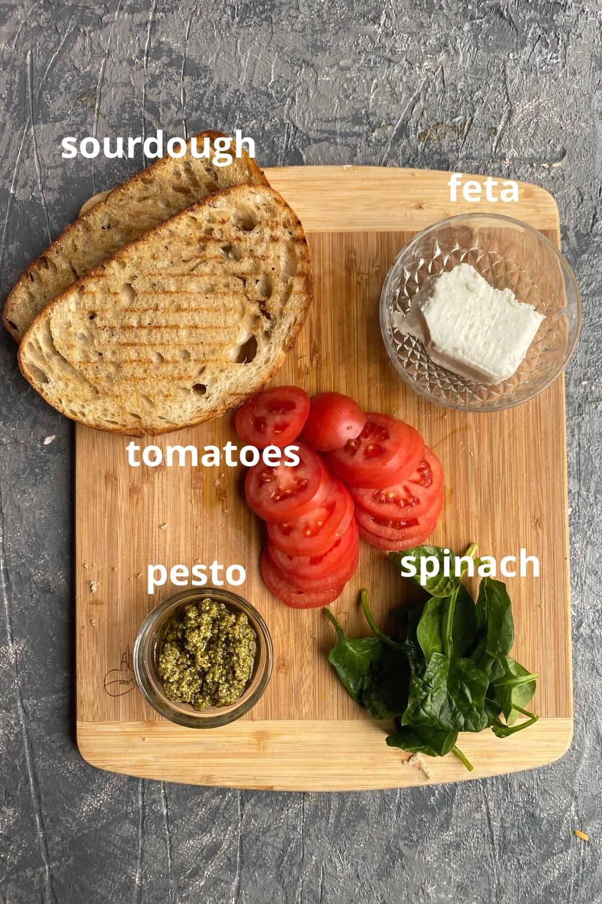 An overhead view of the ingredients to make a grilled cheese sandwich; sourdough, feta, tomatoes, spinach, and pesto