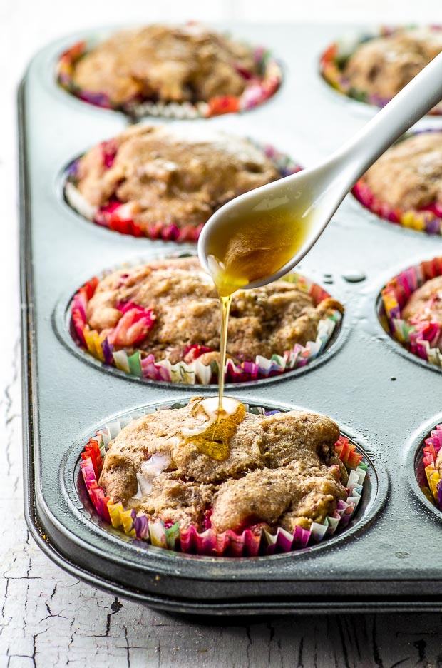 A muffin tin pan with cooked Vegan Pan Roasted Strawberry & Fig Breakfast Muffins. The front muffin shows a white ceramic spoon drizzling some maple syrup on the muffin