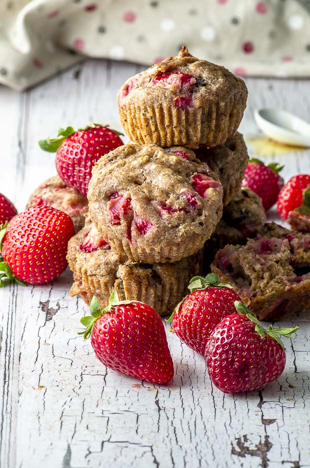 Five Vegan Pan Roasted Strawberry & Fig Breakfast Muffins piled up on a white wood surface. around the muffins there are fresh strawberries and in the background there is a beige napkin with pink, red and white polka dots