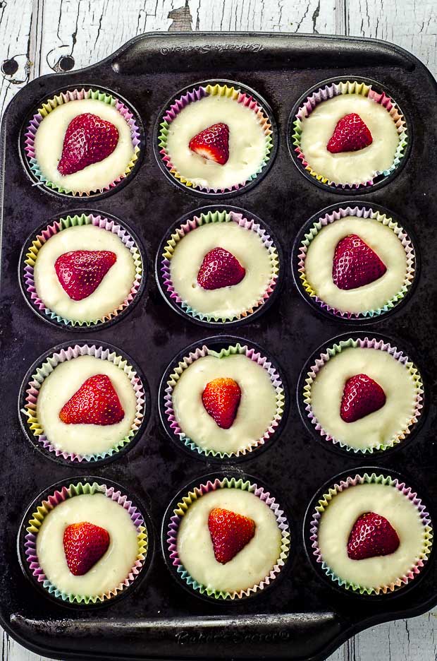 A muffin tray with strawberry muffin batter with ½ a strawberry on top ready to be baked