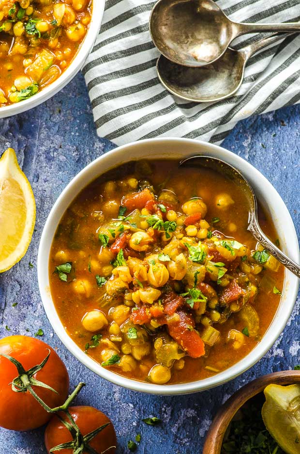 Birds eye view of a white bowl with wheat berry and chickpea soup. On the top left corner there is partial view of another white bowl of chickpea soup. On the right hand corner there is a black and white stripe cloth napkin with 2 spoons on top. On the left side of the chickpea soup bowl there is a slice of lemon. On the bottom left corner there are two tomatoes on the vine.