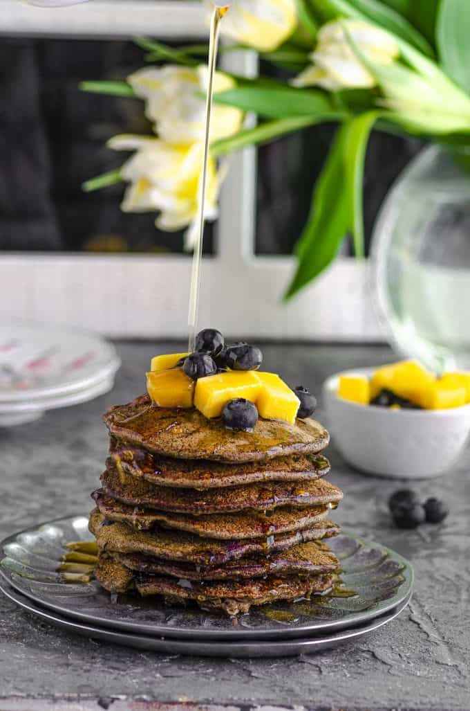 drizzling maple syrup onto buckwheat pancakes topped with mangoes and blueberries