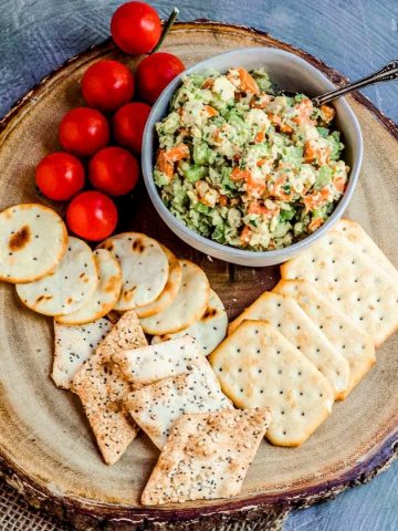 An overhead view of a wood round with a bowl of chickpea tuna salad, various crackers, and cherry tomatoes