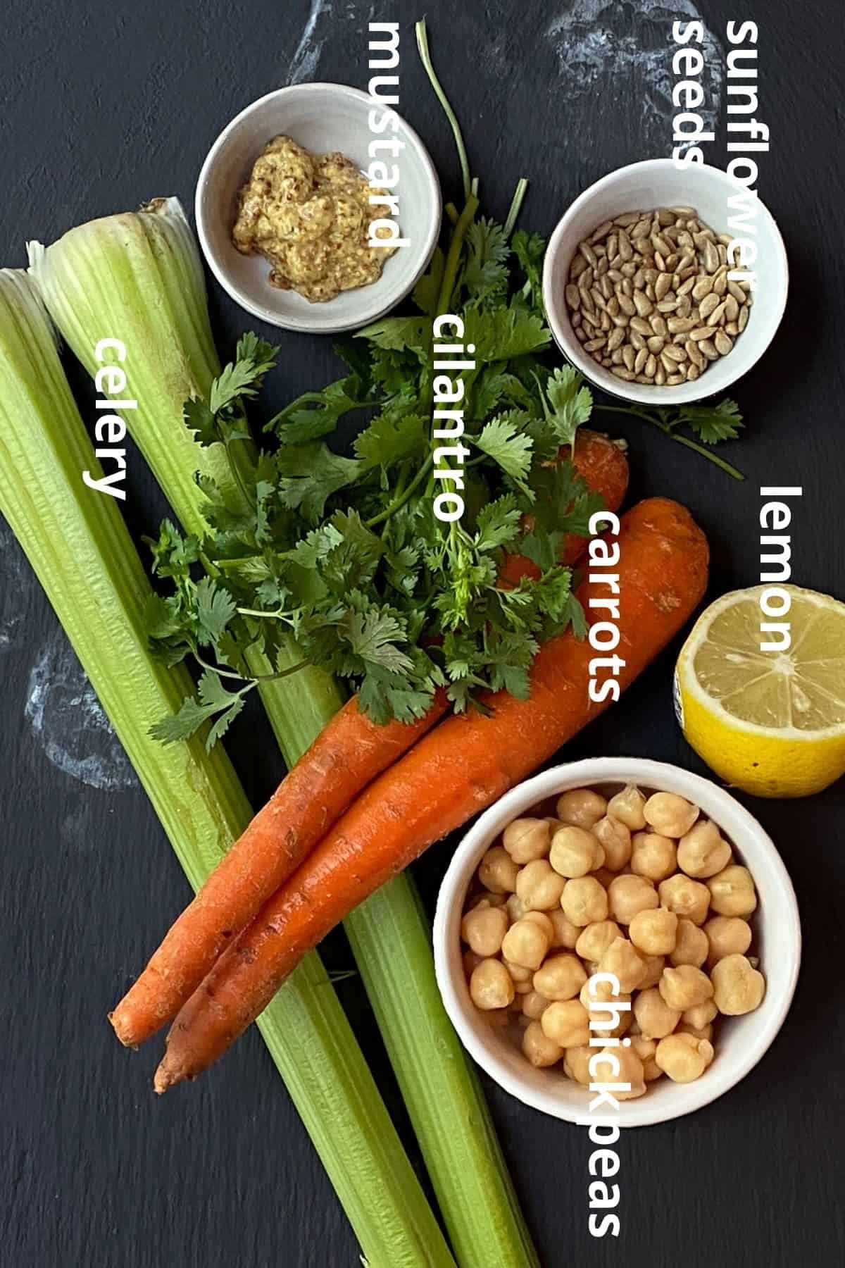 An overhead view of the ingredients to make chickpea tuna salad; sunflower sees, half of lemon, chickpeas, carrots, cilantro, celery, and mustard