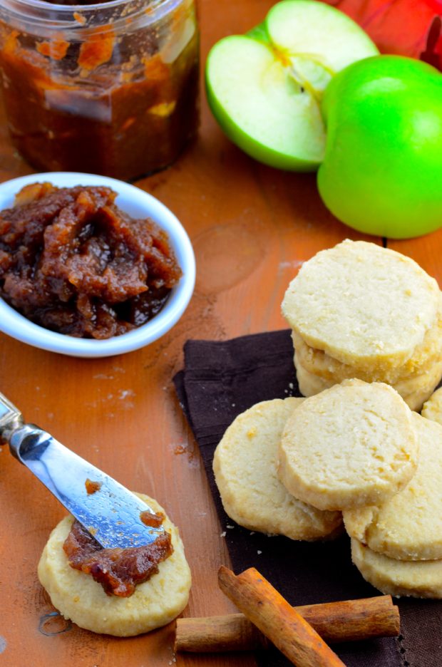 Seven vegan shortbread cookies stacked on a brown place mat. One cookie with a dollop of apple butter spread out on top with a butter knife, and two cinnamon sticks next to it. In the background there is an green apple sliced in half, a half full jar of apple butter, and a small white bowl filled with apple butter