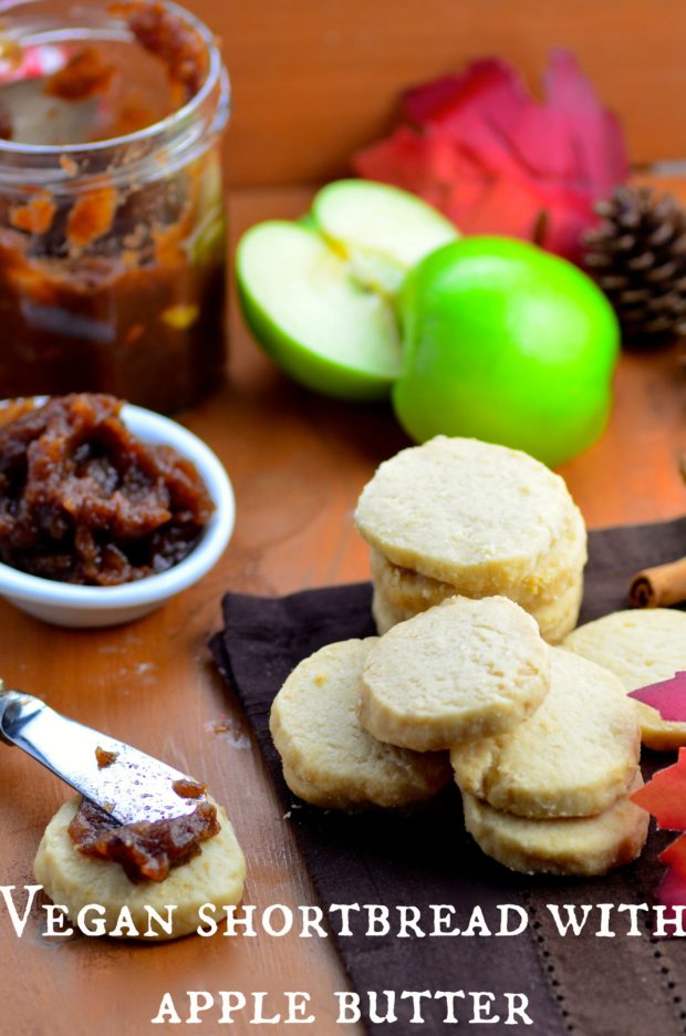 Seven vegan shortbread cookies stacked on a brown place mat. One cookie with a dollop of apple butter spread out on top with a butter knife. In the background there is an green apple sliced in half, a half full jar of apple butter, and a small white bowl filled with apple butter