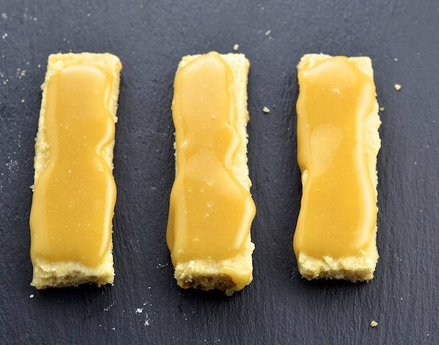 Three shortbread cookies topped with caramel on a dark grey surface