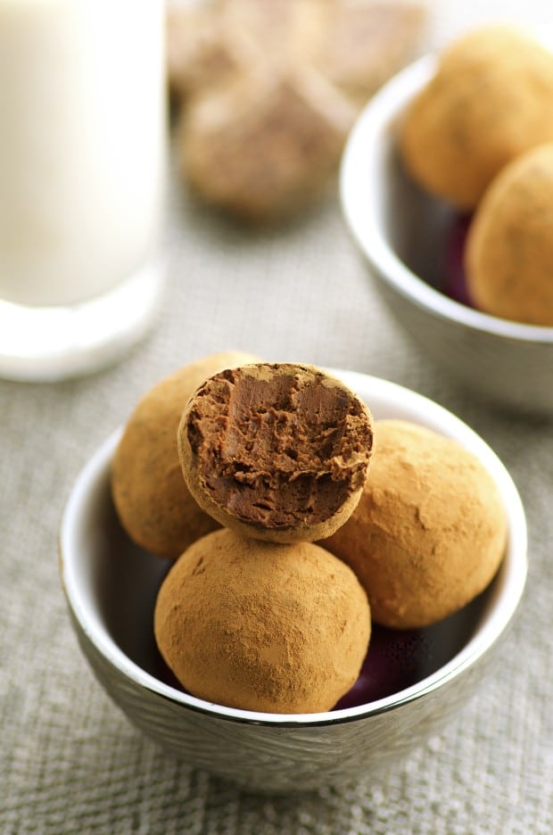 A White bowl filled with Vegan Chocolate Truffles. In the background there is another white bowls filled with vegan chocolate truffles and a glass of milk.