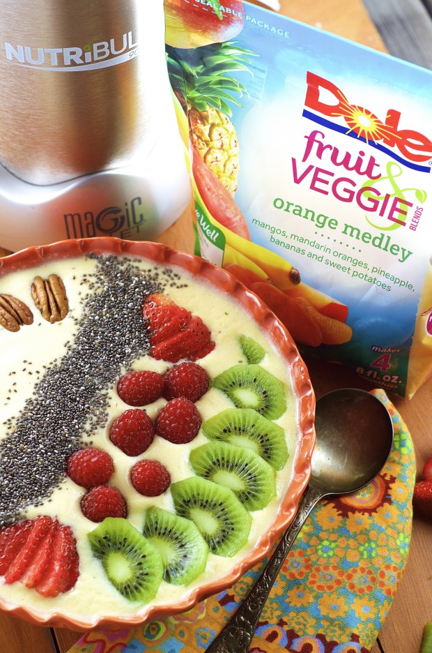 A smoothie bowl with sliced kiwis, fresh raspberries, strawberries, chia seeds and pecans.  In the background there's a pack of dole frozen fruit and a  magic bullet blender.
