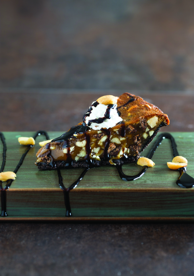 a slice of chocolate chip peanut pie with chocolate drizzle on a green wood plank