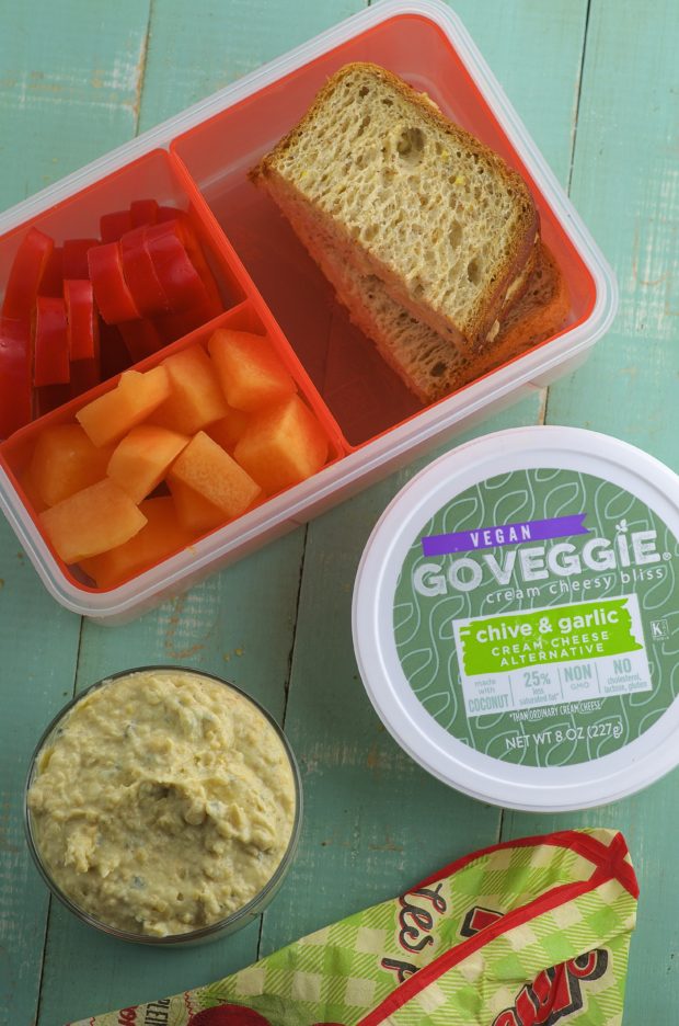 bird's eye view of a lunch box with cut fruit and a chickpea spread sandwich next to a tub of go veggie cream cheese  and a bowl with chickpea spread