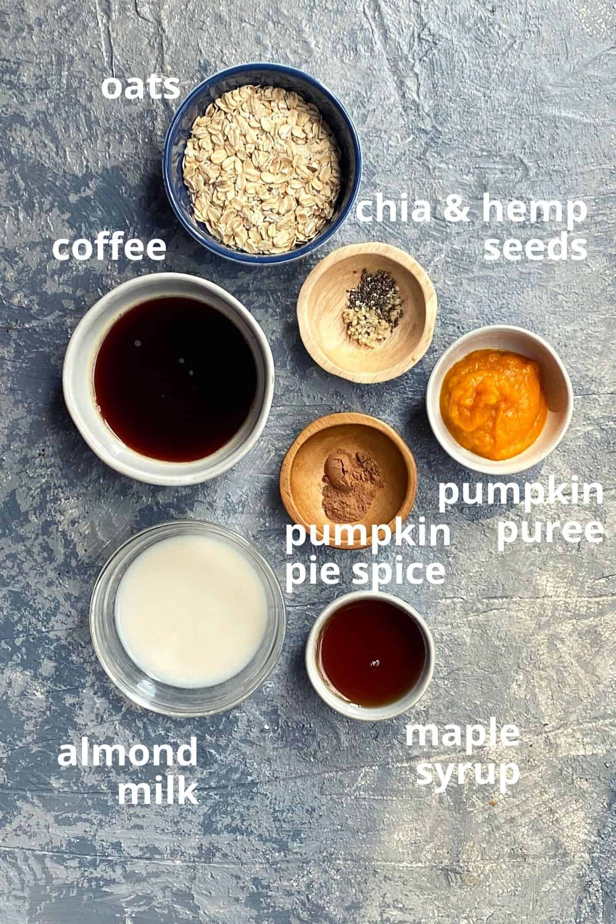 An overhead view of ingredients to make pumpkin spice latte overnight oats in little bowls; oats, chia & hemp seeds, coffee, pumpkin puree, pumpkin pie spice, almond milk, and maple syrup