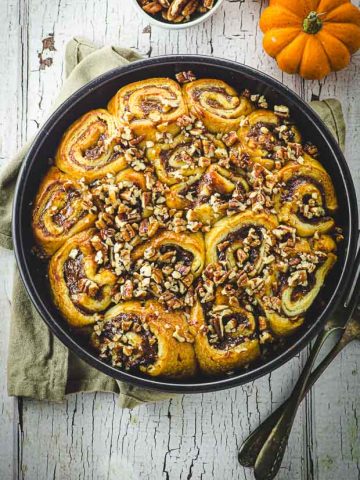 bird's eye view of a round pan filled with baked pumpkin cinnamon rolls . There are two mini pumpkins and a bowl of pecans next to the baking pan.