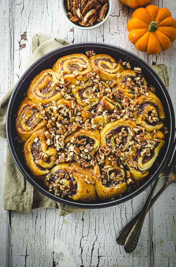 bird's eye view of a round pan filled with baked pumpkin cinnamon rolls . There are two mini pumpkins and a bowl of pecans next to the baking pan.