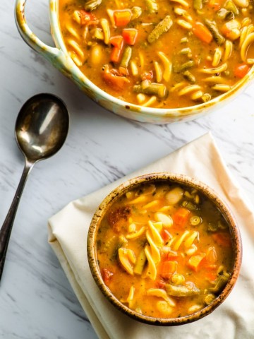 One bowl of minestrone soup next to a serving bowl with soup
