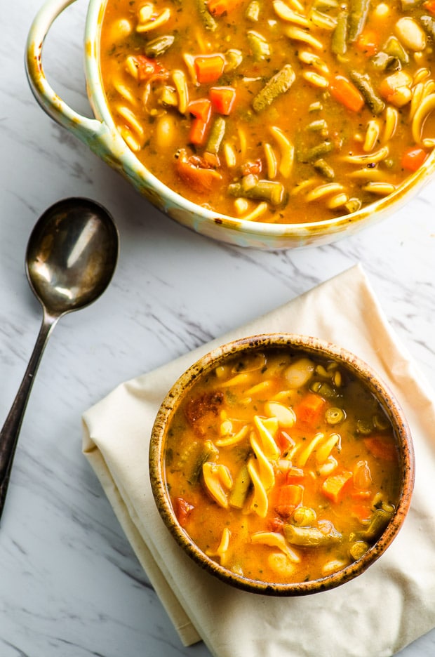 One bowl of minestrone soup next to a serving bowl with soup