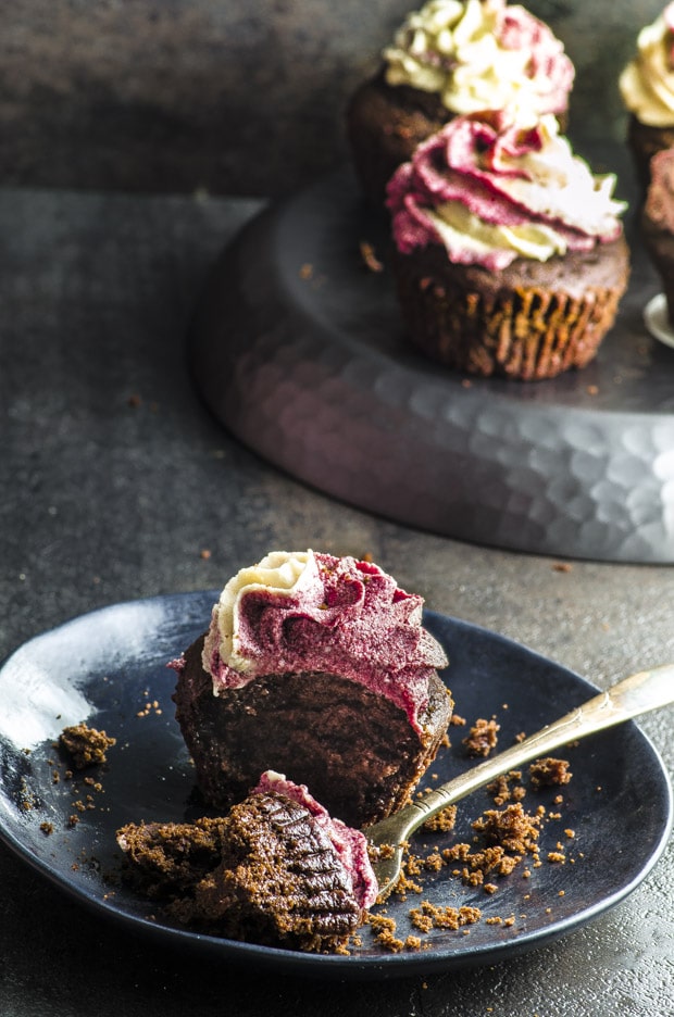 Vegan Pomegranate Tahini Cupcakes - rich and moist chocolate cake topped with creamy tahini and pomegranate molasses icing set on a black ceramic plate. On the background there is a metal plate set upside down with more vegan cupcakes on it. 