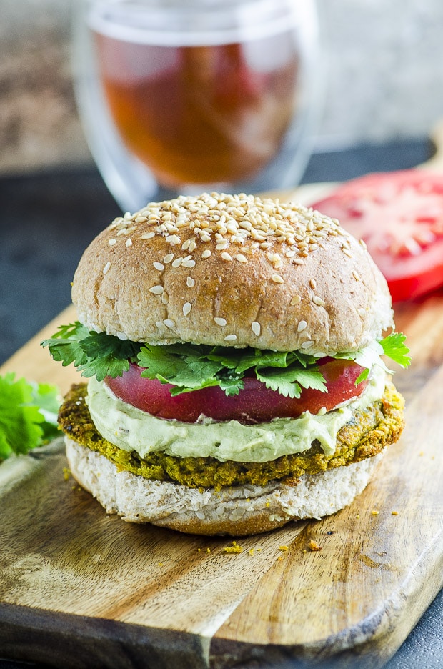 Falafel Veggie Burger With Avocado Tahini Cream on a bun, with a tomato slice and some parley leaves on a wooden cutting board