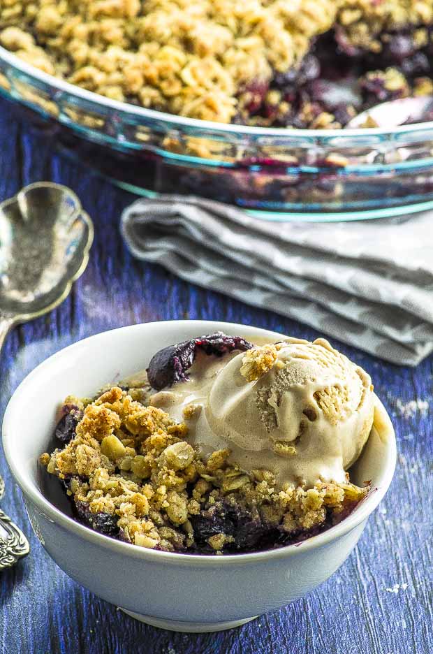 A while bowl with blueberry crisp topped with vanilla ice cream, and with a glass serving dish  in the background
