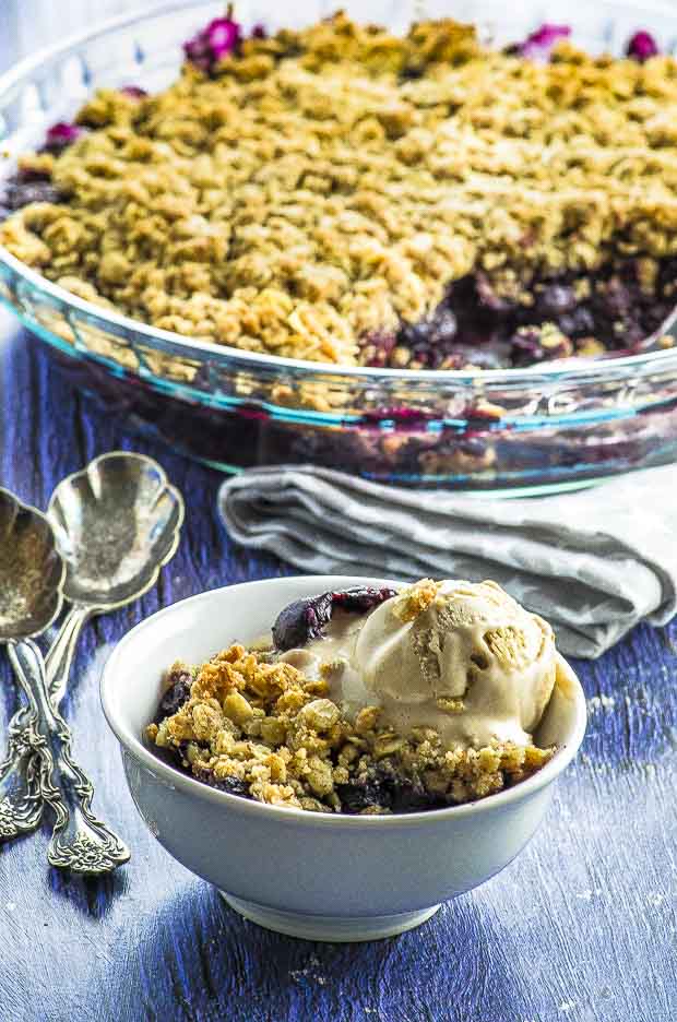 A bowl with blueberry crips a scoop of vanilla ice cream next to a round serving dish with blueberry crumble
