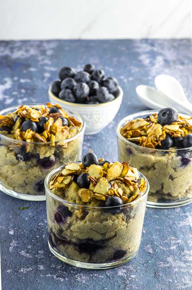 Close up of three clear glass cups filled with blueberry muffin protein grits topped with sliced almonds and a small white bowl of blueberries in the background