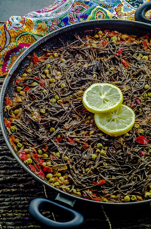This High Protein Noodle Paella is a vegan version of a popular Spanish dish called “Fideua”, traditionally made with fish and shellfish. Vegetarian Black Bean Noodles in a paella pan with vegetables with a couple of slices of lemon.