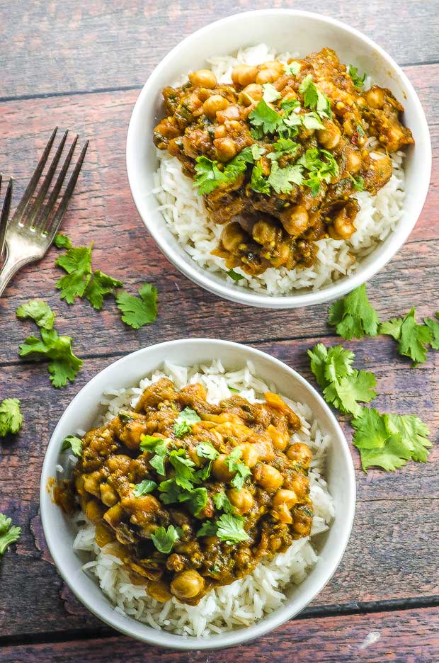 Birds eye view of two white bowls filled with white rice and eggplant and chickpea curry, granished with fresh cilantro, on a wooden surface with a few cilantro leaves scattered on the sides and un fork