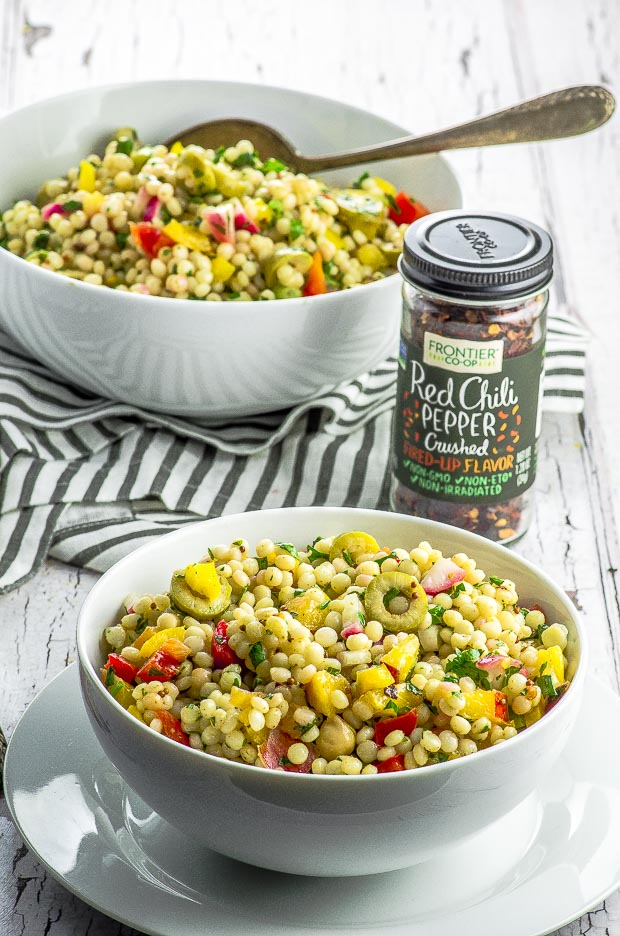 Israeli Couscous Salad with sliced olives, red and yellow peppers and cilantro, in two white bowls, on a white and gray stiped napkin with a small jar of Frontier brand crushed red chili pepper