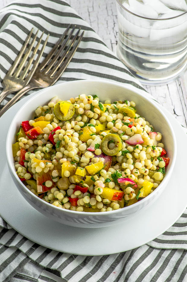 Israeli Couscous Salad with sliced olives, red and yellow peppers and cilantro, in a white bowl, on a white and gray stiped napkin with a glass of ice water and two forks on the side