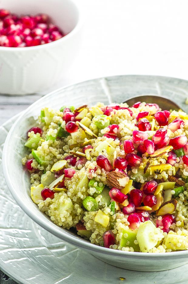 A light grey bowl with quinoa pomegranate salad over a light grey plate, and a small white bowl of pomegranate seeds in the background