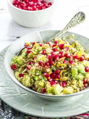 A light grey bowl with quinoa pomegranate salad, with an ornate metal spoon and a small white bowl with pomegranate seeds in the background
