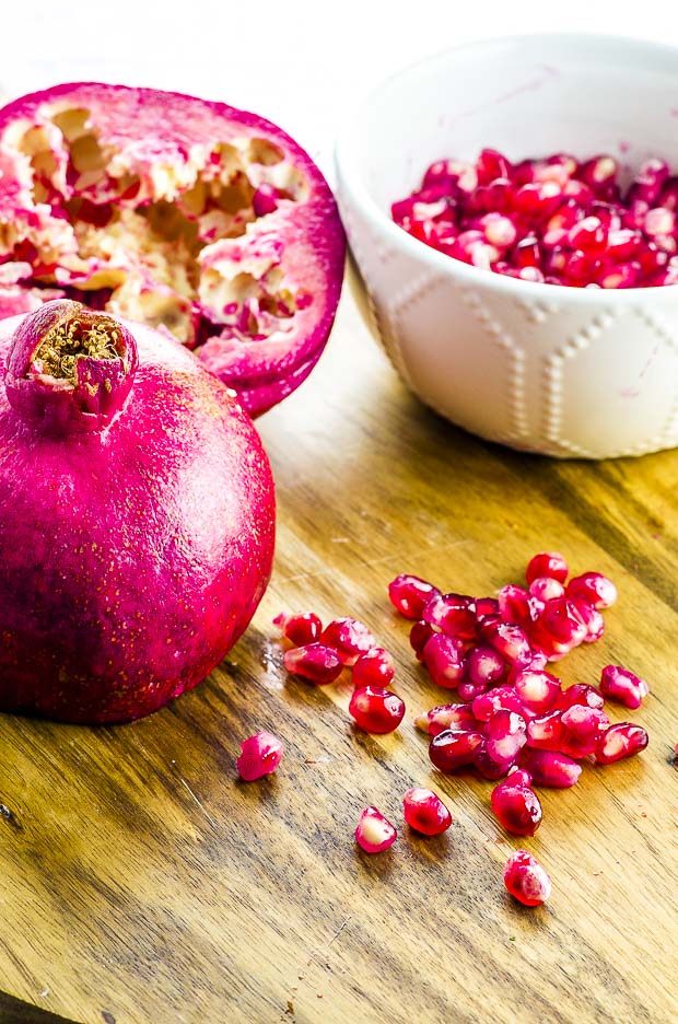 A pomegranate cut in half with the seeds removed on a wooden surface, a few pomegranate seeds scathered next to it and a small white bowl with pomegranate seeds in the background