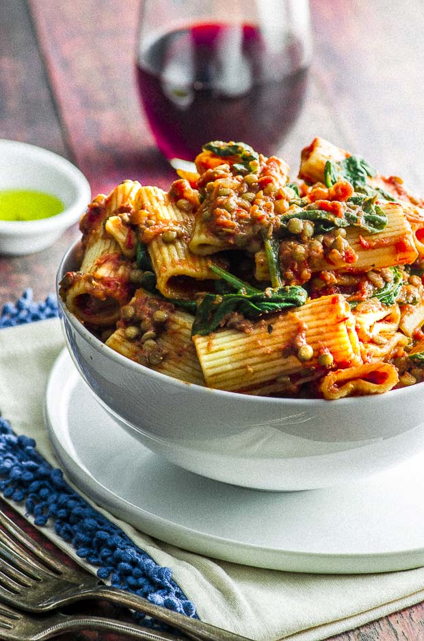 A close up view of a white bowl over filled with rigatoni pasta topped with an easy lentil bolognese and a few baby spinach leaves . The bowl is placed on top of a white plate, under the plate there is a white napkin with a blue fringe. In the background there is a glass of wine, and a small oval white bowl with olive oil.
