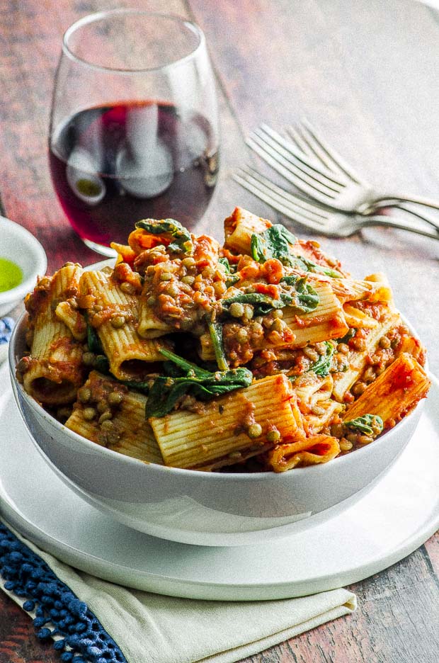 A white bowl over filled with rigatoni pasta topped with an easy lentil bolognese and a few baby spinach leaves . The bowl in placed on top of a white plate, under the plate there is a white napkin with a blue fringe. on the background there is a glass of wine, 3 forks and a sliver view of a small white bowl with olive oil.
