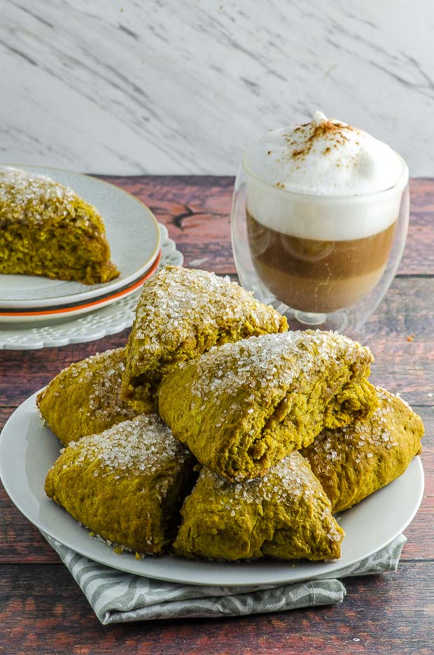 A pile of pumpkin scones on a white plate with a cup of cappuccino in the background
