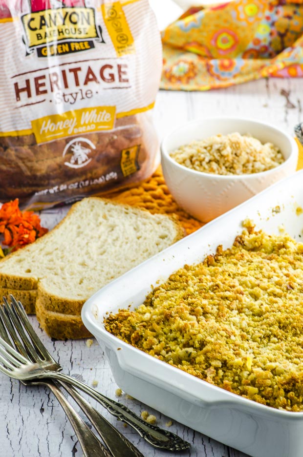 Gluten free butternut squash and cauliflower gratin in a white baking dish, with two forks and two slices of bread placed on the left side. In the background there is a package of Canyon Bakehouse bread and a small white bowl with bread crumbs