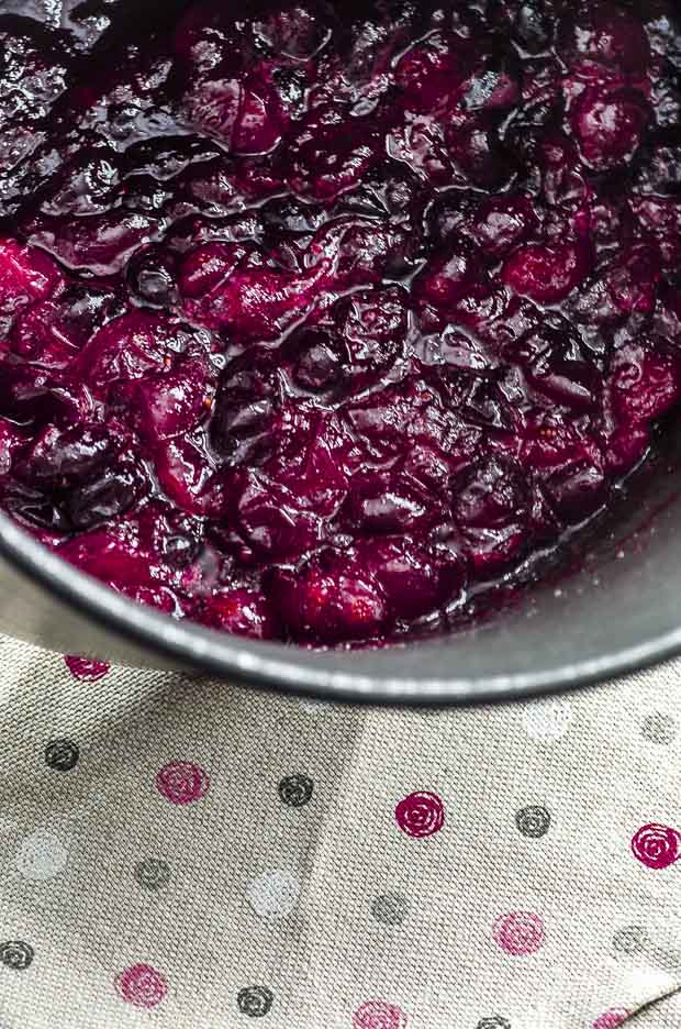Close up of cooked cranberries in a saucepan over a gray cloth napkins with red, white and gray polkadots