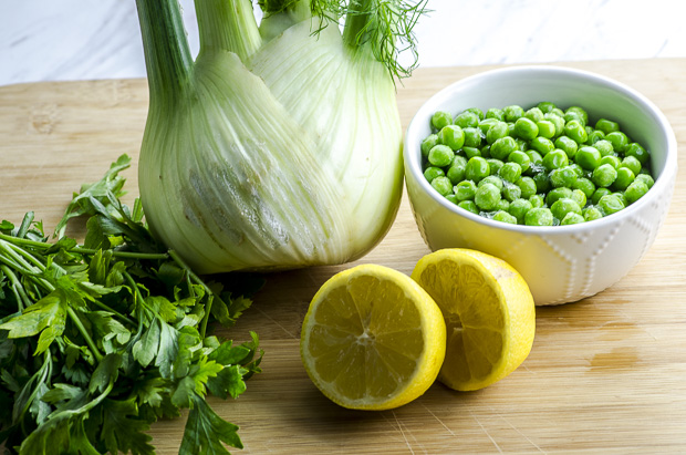 A fennel bulb, a bunch of parsley, a white bowl with frozen peas and a lemon cut in half on a wooded cutting board 